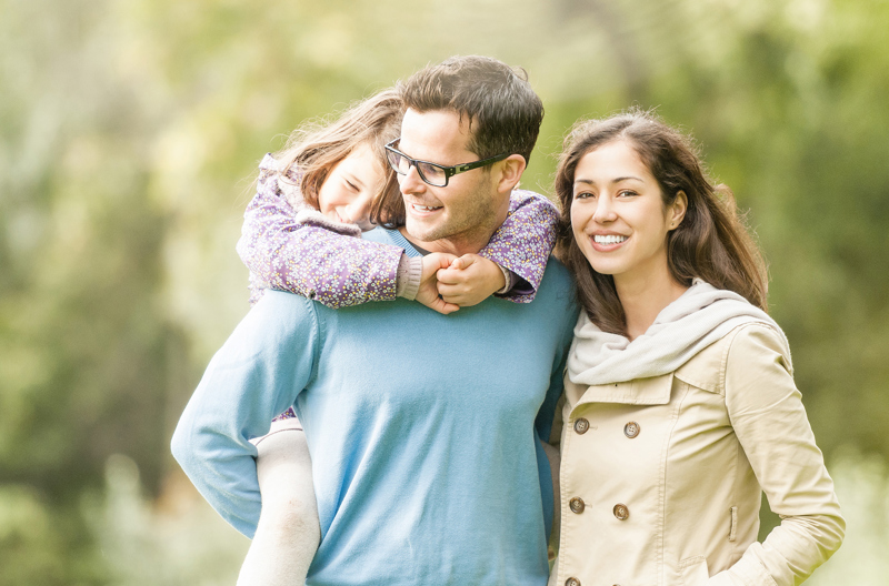 Two parents standing in a park with one of the parents holding their child on their back.