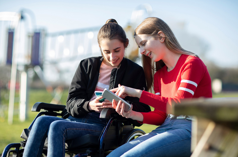 Two friends sitting together at a park showing each other something on their phones.