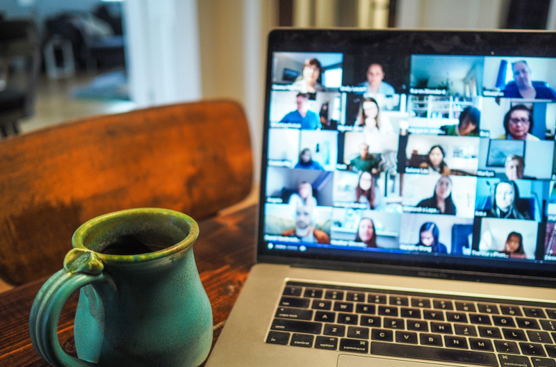 A cup of coffee on a table next to an open laptop with a group video call showing. 