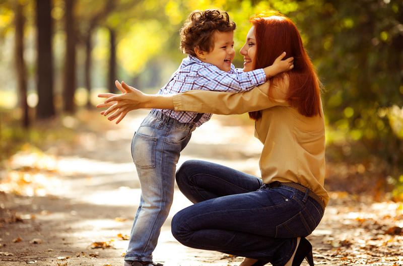 A mother kneeling down at the park to hug her child. 