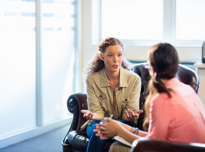 Two people sitting on separate couches talking while one person takes notes. 