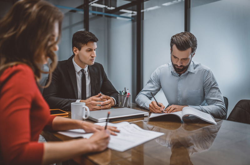 Three people wearing business attire writing on contracts in an office. 