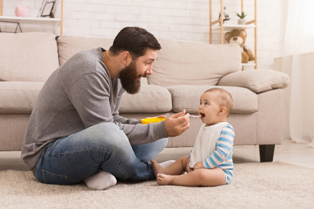 A young dad sitting on his living room carpet, feeding his infant child with a spoon.