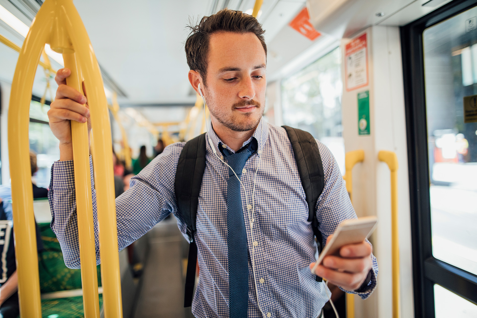 A man wearing a shirt and tie wearing earphones attached to a phone as he stands on a tram.