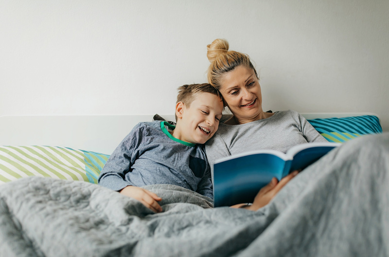 A parent laying in a bed with her child reading a book. 