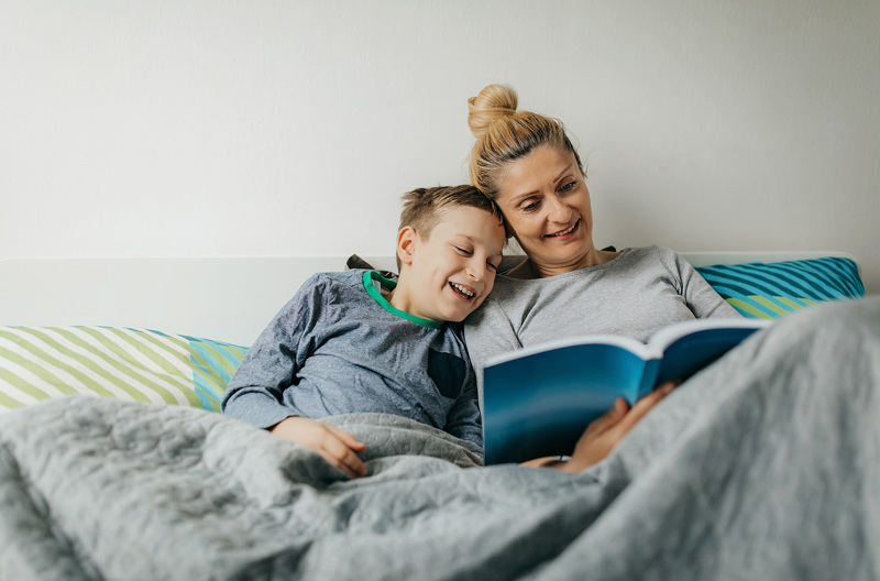 A parent laying in a bed with her child reading a book. 