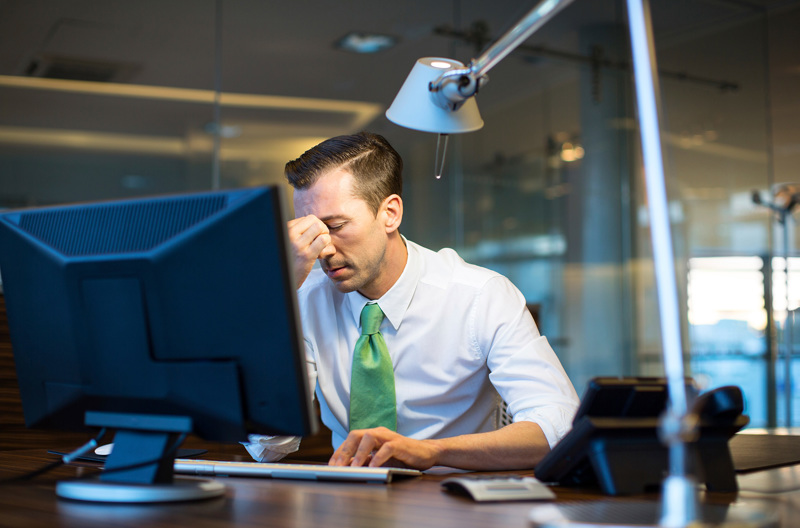 A man wearing a white shirt with a green tie sitting at a desk with a disgruntled look on his face.