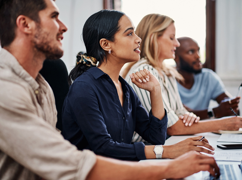 Four people sitting next to each other at a table writing notes on their notepads or laptops