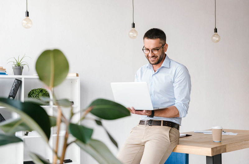 A man wearing a white business shirt leaning against a wooden table as he holds an open laptop.