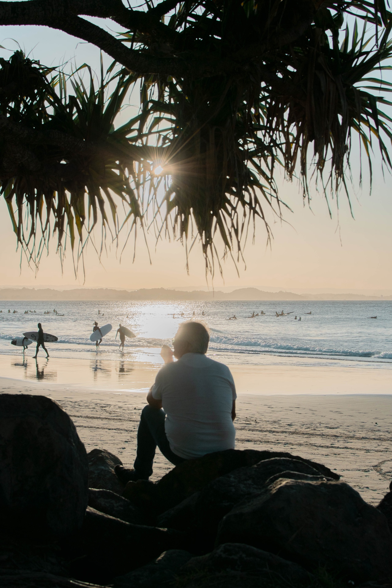 A man sitting on a rock at the beach as the sun sets in the background.