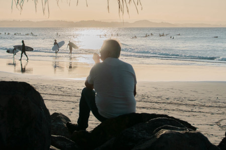 A man sitting on a rock at the beach as the sun sets in the background.