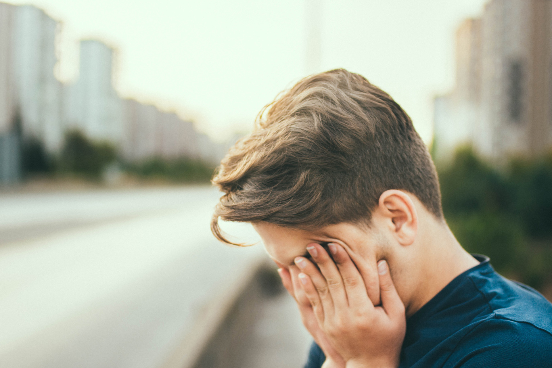 A man standing outside next to a road covering his face with his hands.