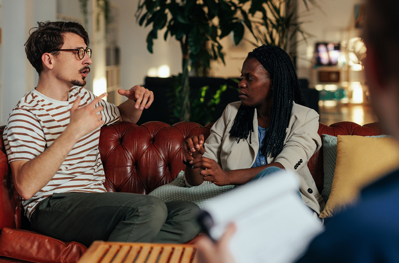 A biracial couple sitting on a couch and having a serious conversation while a couples counsellor takes notes