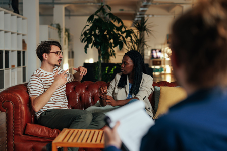 A biracial couple sitting on a couch and having a serious conversation while a couples counsellor takes notes 