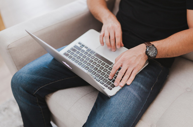 A man sitting on a couch with a grey laptop on his lap.