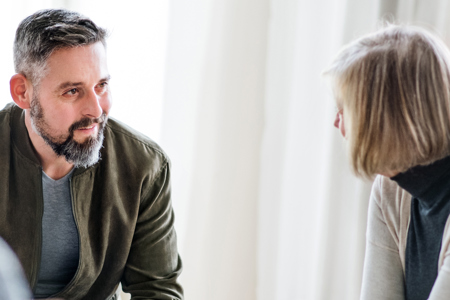 A man and a woman, seated indoors, having a friendly conversation. The man is Caucasian, with short, dark hair and a beard and wears a grey t-shirt under a khaki green jacket. The women is Caucasian with a shoulder length blond bob, and wears a dark, turtleneck top and a cream cardigan.