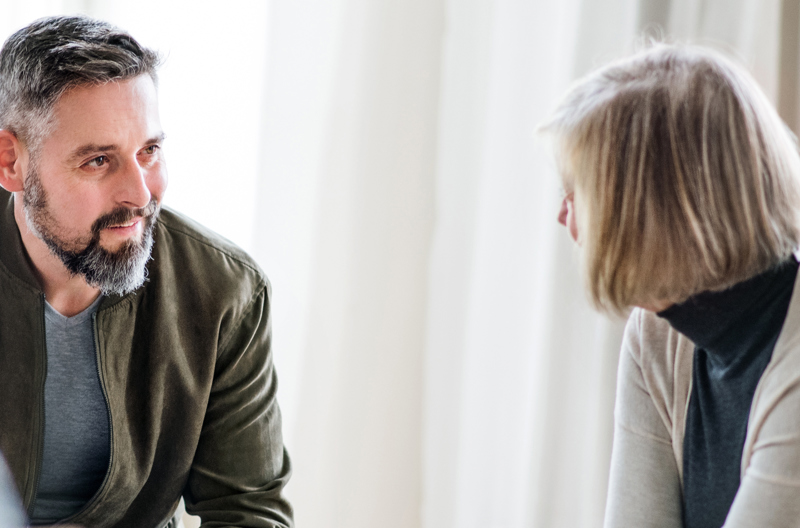 A man and a woman, seated indoors, having a friendly conversation. The man is Caucasian, with short, dark hair and a beard and wears a grey t-shirt under a khaki green jacket. The women is Caucasian with a shoulder length blond bob, and wears a dark, turtleneck top and a cream cardigan.