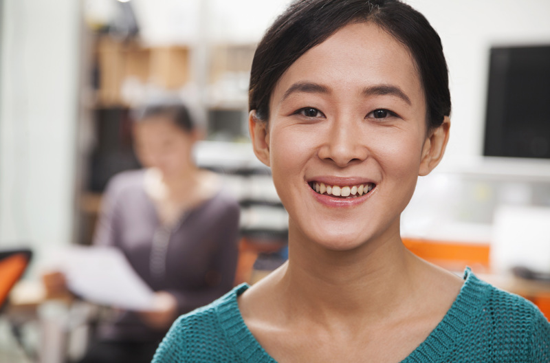 A young woman wearing a green top smiling at the camera while someone reads off a piece of paper in the background.