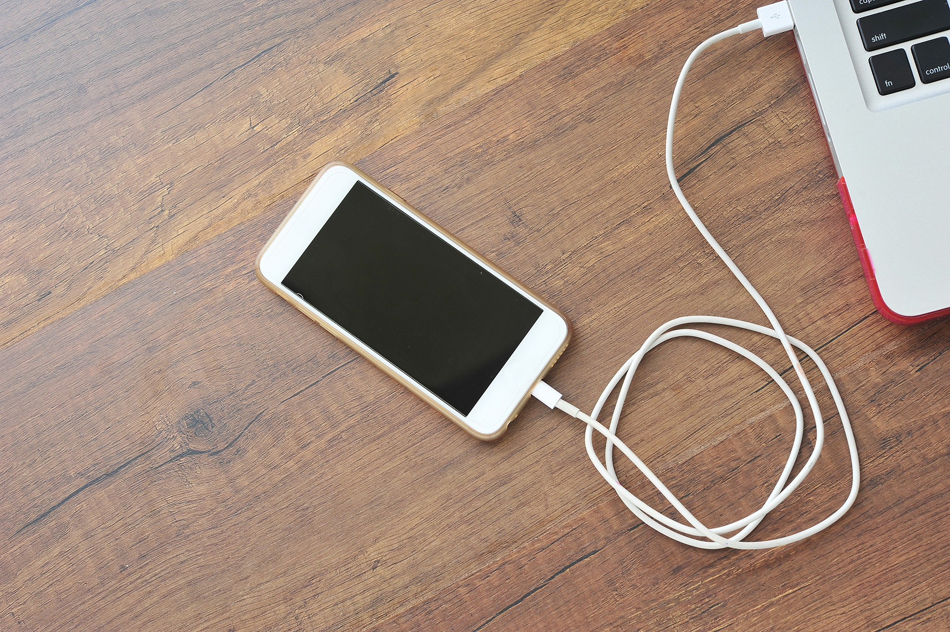 A smart phone connected by a cable to a laptop, sitting on a wooden table.