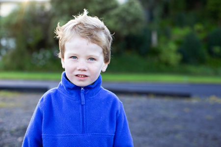 A young boy wearing a blue zip sweater standing in front of trees.